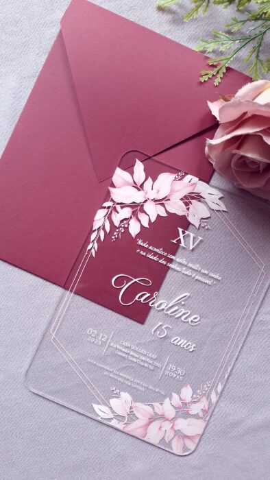 Convite de 15 Anos em Acrílico com Impressão Floral e Envelope Mymalmero | Art Invitte 1 Convite de 15 anos de luxo feito em acrílico transparente com bordas arredondadas, impressão de flores rosas e texto em serigrafia branca, posicionado sobre um envelope de papel Mymalmero Palissandre cor marsala.